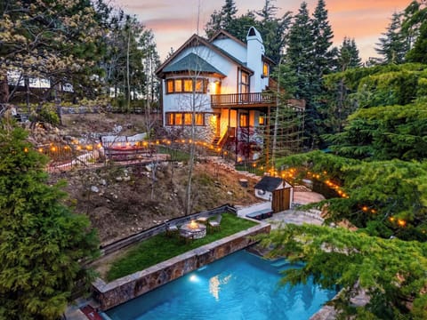 Warm mountain retreat with a glowing pool, fire‑pit lounge, and sunset views framed by evergreens.