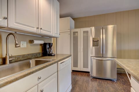 A modern kitchen with white cabinets, a stainless steel refrigerator, a sink, a dishwasher, and a coffee maker on a countertop. The floor is wooden, and the walls have vertical paneling.