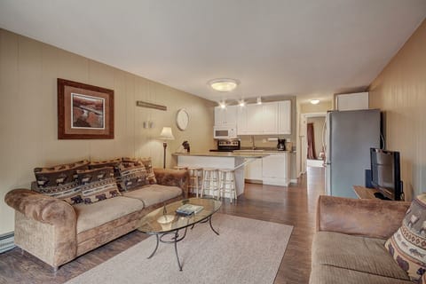 A cozy living room with a brown sofa, patterned pillows, a glass coffee table, and a TV. It is adjacent to a kitchen area with white cabinets, an island with two stools, and various appliances.