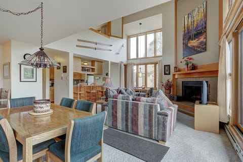 Spacious living and dining area with high ceilings, striped couches, a fireplace, large windows, and an open kitchen in the background.