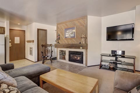 A cozy living room with a fireplace, a mounted TV, a wooden rocking chair, and a gray sectional sofa. A wooden coffee table sits in front of the sofa. The kitchen area is visible in the background.