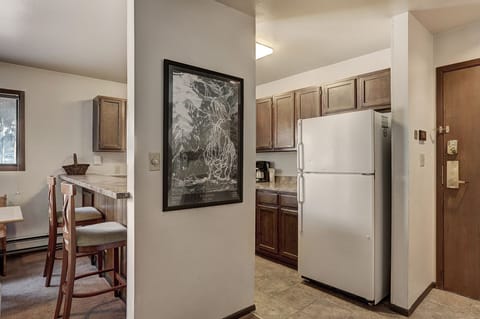 A kitchen and dining area with wooden cabinets, a white refrigerator, bar stools at a counter, a dining table with chairs, and a framed picture on the wall.