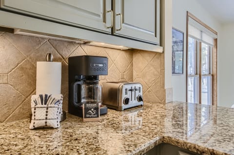 A kitchen countertop with a coffee maker, toaster, paper towel roll, and cloth towels near upper cabinets and a window.