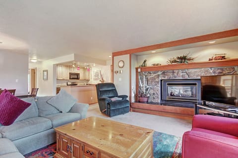 A cozy living room with a stone fireplace, gray sectional sofa, red armchair, wooden coffee table, and an open view of the kitchen area in the background.
