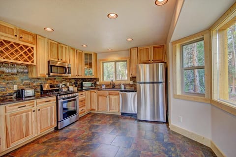 A modern kitchen with wooden cabinets, stainless steel appliances, a tiled floor, and large windows overlooking greenery.