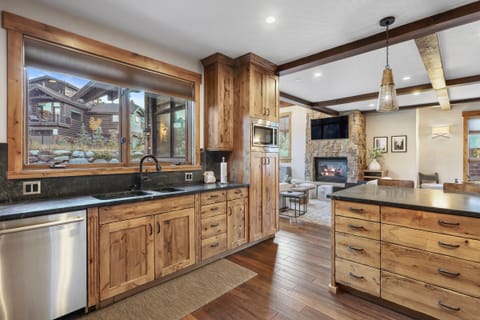 A modern kitchen with wooden cabinets, granite countertops, stainless steel appliances, and an island with drawers. The kitchen opens into a living area with a dining room.