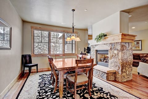 A dining room with a rectangular wooden table, four chairs, a light fixture above, a large window, and a stone fireplace with a mantel in a cozy interior space.