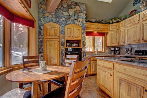 A rustic kitchen-dining area with wooden cabinets, a round table with four chairs, a stone wall, appliances, and a window with a red valance overlooking a snowy outdoor scene.