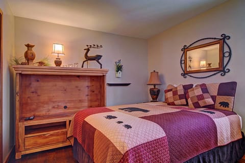 A cozy bedroom with a quilt featuring bear and moose designs on a double bed, a wooden cabinet with a mounted deer figure, and a mirror in a decorative frame above a lamp-lit nightstand.