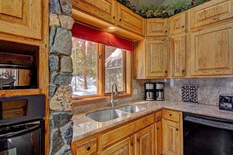 A rustic kitchen with wooden cabinets, a stone pillar, stainless steel sink, and modern appliances. A window above the sink shows a snowy outdoor scene. Potted plants are placed above the cabinets.