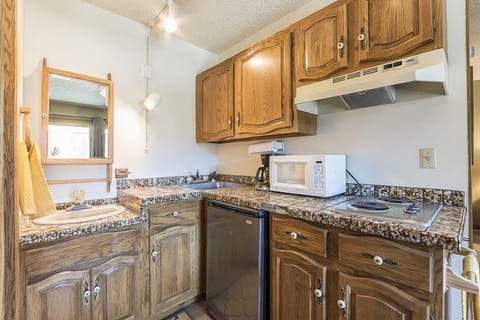A compact kitchen with wooden cabinets, granite countertops, a small sink, a mini-fridge, a microwave, and a two-burner stove. A small wall-mounted mirror and towel are located on the left side.