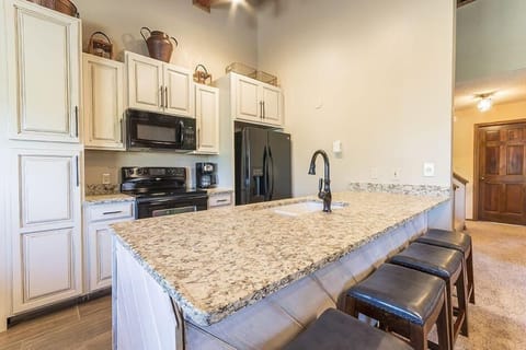 Modern kitchen with granite countertops, white cabinetry, black appliances, and a black faucet. Three black stools are lined up along the island. Light walls and a wooden door are in the background.