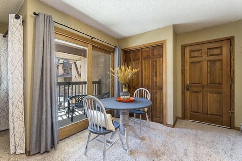 A small dining area features a round table with two chairs, a vase with dried plants, and a sliding glass door with a balcony view. The room has carpeted floors and wooden details on the doors and cabinets.