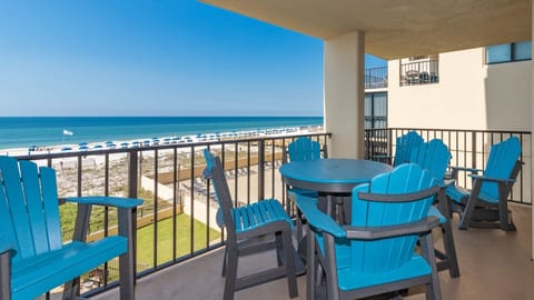 The balcony features a round table surrounded by vibrant blue chairs, offering a view of the beach and umbrellas