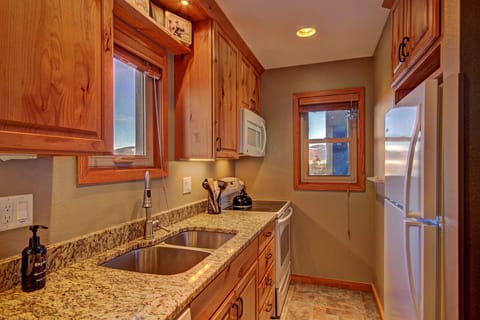 A compact kitchen with wooden cabinets, granite countertops, a double sink, refrigerator, microwave, stove, and small appliances on the counter. Natural light enters through two windows.