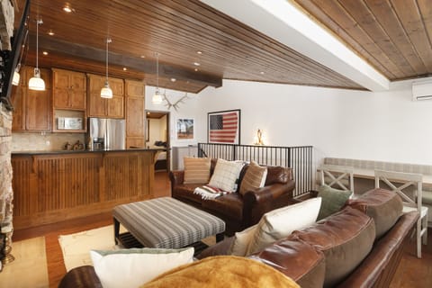A cozy living room with a brown leather sofa, checkered ottoman, and wooden ceiling. The background shows a kitchen area with wooden cabinets and a bar counter. An American flag decorates the wall.