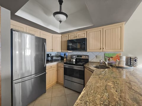 A modern kitchen with light wood cabinets, stainless steel refrigerator, stove, microwave, and granite countertops. The counter has a sink and various kitchen items.