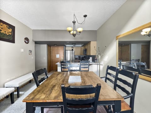 Dining area with a wooden table and six chairs, overlooking a kitchen with stainless steel appliances. A chandelier hangs above the table, and a large mirror is mounted on the wall to the right.
