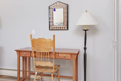 A wooden desk and chair are placed against a white wall with a decorative mirror. A black floor lamp with a white shade stands to the right of the desk.