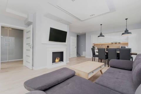 A spacious living room with a cozy sectional sofa, a wooden coffee table, a large wall clock, and abundant natural light coming through the sliding glass doors.