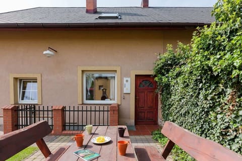 View of the house from the front. A wooden table with benches is visible in the foreground and the entrance to the house can be seen behind them. 