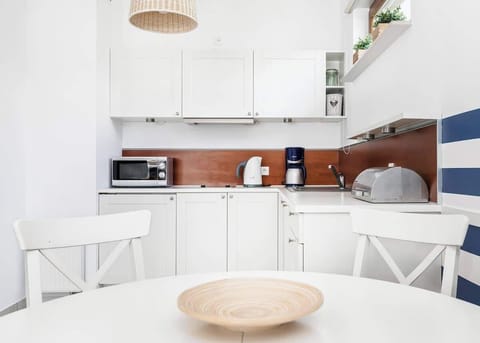 In the foreground, a side table with a decorative bowl can be seen, and behind it is the kitchen area with white furniture, microwave, coffee machine and essential utensils.