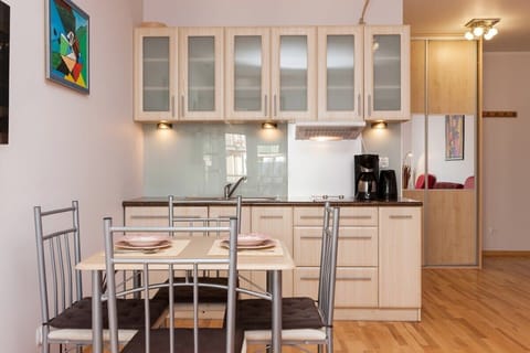 Kitchen connected to the dining room. The room has a light wooden floor and the kitchen cabinets are made of light wood. The dining table is next to the sofa and the whole room is lit with warm light.