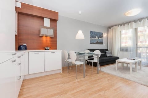 A cozy open-plan living area with a white dining table in the foreground. The kitchen is visible in the background, featuring wooden accents and modern lights.