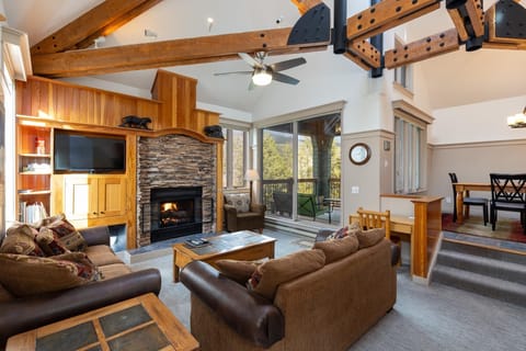 A spacious living room with wooden beams, a stone fireplace, sofas, a television, and large windows overlooking a forest. A dining area with a table and chairs is visible in the background.