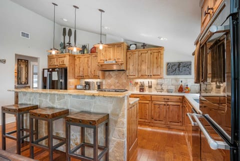 A rustic kitchen features light wood cabinets, stone-clad island with bar stools, and modern appliances, under pendant lights.