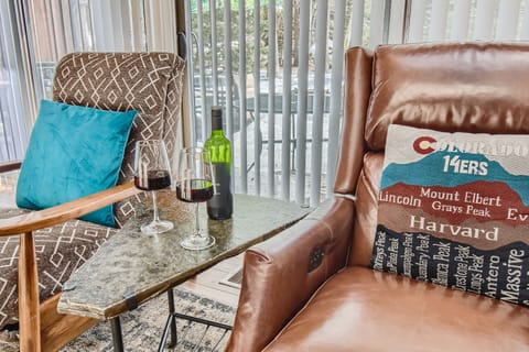 Two chairs with cushions sit beside a table holding two glasses of red wine and a wine bottle. A window with vertical blinds and a view of trees is in the background.