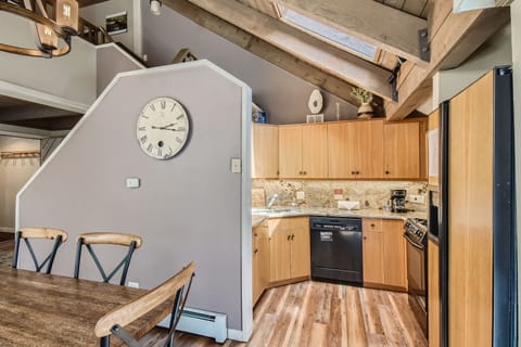 A modern kitchen with wooden cabinets, marble countertops, stainless steel appliances and a skylight above. A large wall clock decorates the adjacent wall. A dining area with a wooden table is visible.