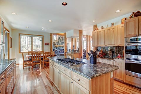 A spacious kitchen and dining area with wooden cabinets, granite countertops, a central island with a stove, and a dining table by the window offering a view of a wooded area.