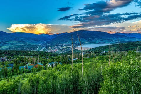 A panoramic view of a lush green valley with scattered houses, a distant lake, and mountains under a cloudy sky at sunset.