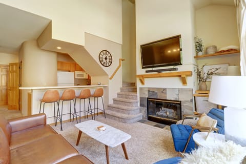 A modern living room with a wall-mounted TV, a fireplace, a leather couch, a marble coffee table, and a kitchen with barstools. A staircase leads to an upper level.