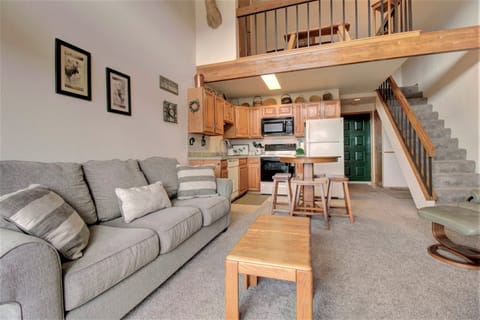 A cozy living room features a grey sofa, coffee table, kitchen with wooden cabinets, black appliances, a dining area, and a staircase leading to an upper level.