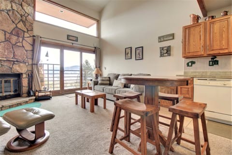 A cozy living room with a stone fireplace, a gray sofa, wooden furniture, a high table with stools, and a view of the mountains through large windows. The kitchen area has wooden cabinets and white appliances.