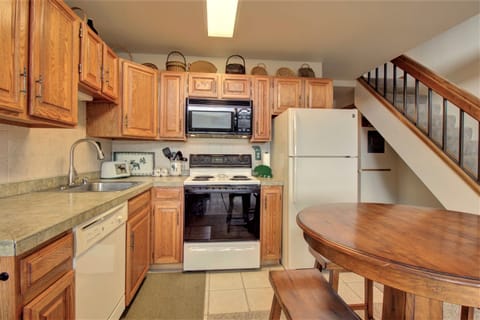 A small kitchen with wooden cabinets, a microwave, stove, and refrigerator. There is a sink, dishwasher, and a round wooden table with chairs. A staircase is visible in the background.