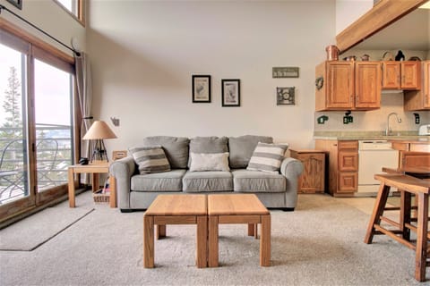 A cozy living room with a gray couch, two wooden coffee tables, a sliding glass door leading to a balcony, and an adjoining kitchen with wooden cabinets.