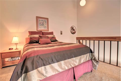 A bedroom with a neatly made bed featuring a striped comforter, a nightstand with a lit lamp, and framed art on the wall. The room has a high ceiling and railing beside the bed.