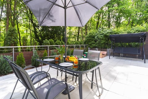 An outdoor dining area with a patio table, four chairs, and a large umbrella. The space is surrounded by greenery, creating a relaxing atmosphere.