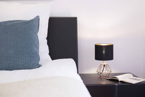 A close-up of a bedside table with a lamp, next to a bed with crisp, white bedding. The lighting creates a warm ambiance.
