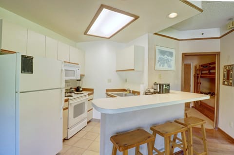 A small kitchen with white appliances, a countertop bar with three wooden stools, and hallway leading to another room with a bunk bed.