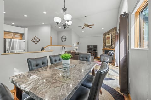 Stylish dining room featuring a polished stone-top table and plush seating for six. Large windows and glass doors frame a peaceful forest backdrop, seamlessly connecting to the cozy living area and stone fireplace.