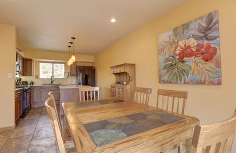 A kitchen and dining area with a wooden table and chairs, pendant lights, a floral wall painting, and cabinets. Kitchen appliances are visible in the background. Walls are painted in a light color.
