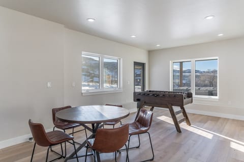A room with a round wooden table and five brown chairs on a light wood floor. There is a foosball table near windows showing an outdoor view.