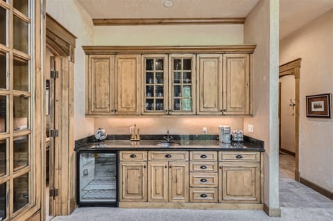A small kitchen area with wooden cabinets, a black granite countertop, a mini-fridge, a stainless steel sink, and various kitchen items, including a coffee maker and utensils.