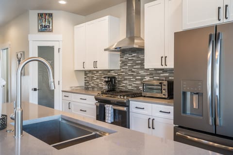 Modern kitchen with white cabinetry, stainless steel appliances, including a double-door fridge and oven, and a gray tiled backsplash. The countertop features a double sink with a high-arc faucet.