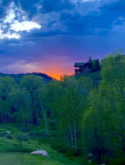 A house on a hill is silhouetted against a vibrant sunset sky with clouds, surrounded by green trees and grassy landscape.
