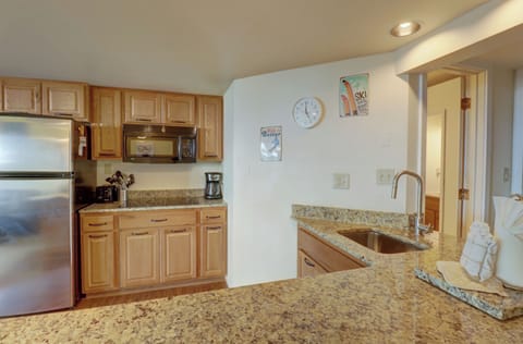 A modern kitchen with granite countertops, stainless steel fridge, light wood cabinetry, sink, coffee maker, and wall clock. Decor includes a "Ski Utah" sign and a "Home Sweet Home" sign.